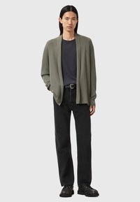 Cárdigan verde oliva con apertura frontal, textura acanalada; usado sobre una camiseta oscura y jeans negros; completado con zapatos negros con cordones.
