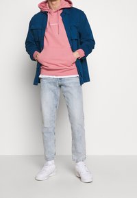 Camicia blu con bottoni sopra una felpa con cappuccio rosa, abbinata a jeans chiari e sneakers bianche. La felpa ha una tasca a marsupio e coulisse.