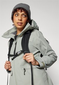 Light grey waterproof jacket with a high collar, adjustable hood, and zippers. Black backpack straps with clips. Knit grey beanie.