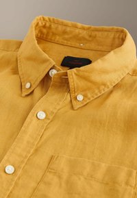 Chemise en lin jaune moutarde avec col, boutons blancs et poche poitrine, affichant une étiquette "Next Signature" à l'intérieur du col.