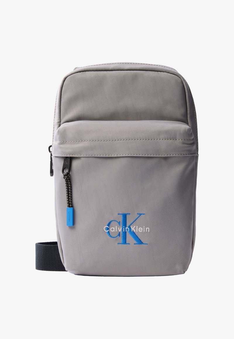 Μικρή γκρι τσάντα Calvin Klein για να φοριέται χιαστί, με μπροστινή τσέπη με φερμουάρ, μαύρο λουρί και κεντημένο μπλε λογότυπο "cK".