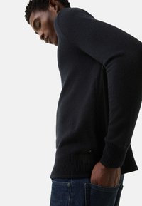 Marineblauer Strickpullover mit gerippten Bündchen und Saum. Weiche Textur und tailliertes Design. In dunkle Jeans gesteckt, Hand in der Tasche.