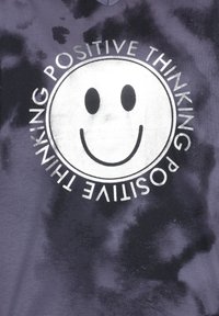 Violetti solumuotokuvio t-paita, jossa on valkoinen pyöreä graafinen kuva, jossa on hymyilevä naama ja sanat "POSITIIVINEN AJATTELU" voimakkailla mustilla kirjaimilla.