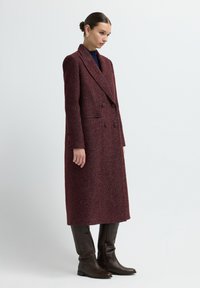 Mujer vestida con un largo abrigo de espiga en color marrón burdeos de doble botonadura y botas de cuero marrón oscuro hasta la rodilla, de pie contra un fondo liso.