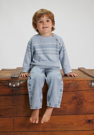 Hellblauer Pullover mit einem gemusterten Design, drei dekorativen Knöpfen und hellblauen Jeans. Kind sitzt barfuß auf einem Holztrunk.