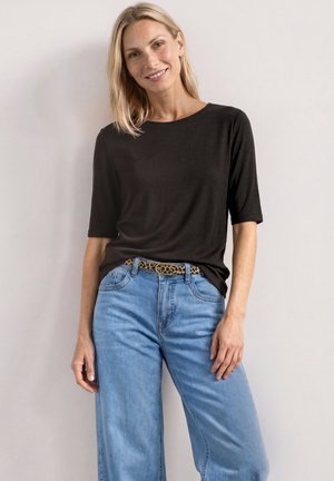 Lyshåret kvinde smilende iført en sort top med korte ærmer, lyseblå jeans og et leopardmønstret bælte, der står op ad en ensfarvet væg.