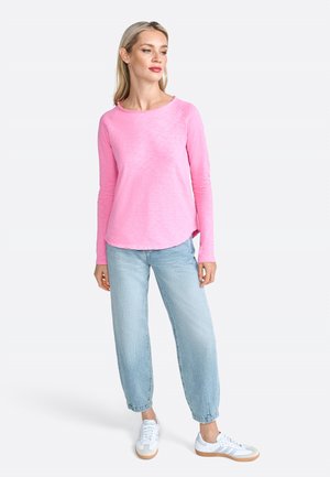 Camicia rosa chiaro a maniche lunghe con scollatura rotonda e orlo leggermente curvato, abbinata a pantaloni larghi azzurri e sneakers bianche.
