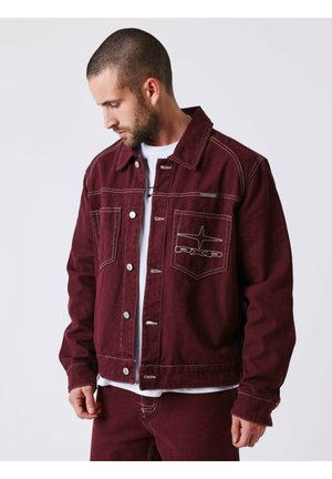 Homme portant une veste bordeaux boutonnée et un pantalon assorti, regardant vers le bas à sa gauche sur un fond uni.