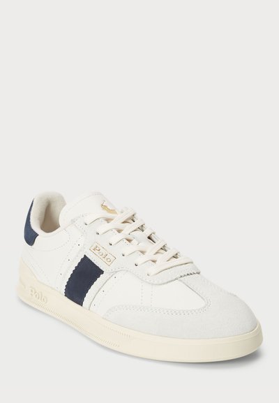 White sneaker with navy accents, suede and leather materials, round toe, flat cream sole, and lace-up design featuring a logo detail.
