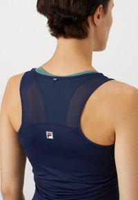Marineblå atletisk tank top med mesh bakpanel, grønn trim og en liten logo i midten. Glatt, tettsittende tekstur.