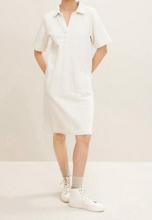 Abito bianco a maniche corte con colletto e scollatura a V, dotato di due tasche laterali. Indossato con sneakers bianche alte e calzini grigio chiaro.
