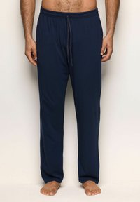 Man med marinblå dra på joggingsbyxor, stående barfota mot en enkel ljus bakgrund.