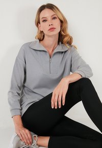 Siv pulover s zapenjanjem na ovratniku, z teksturirano sprednjo ploščo in dolgimi rokavi, v kombinaciji s črnimi legicami in sivimi športnimi copati.