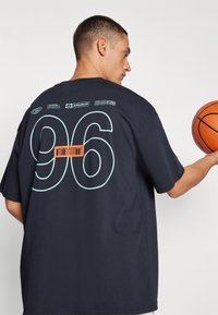 Černé oversized tričko s velkým světle modrým "96" na zádech, s oranžovým akcentovým textem a bílými a modrými logy tištěnými nad ním.