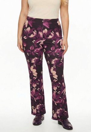 Kvinde iført lilla bukser med blomsterprint, beige top og mørkelilla loafers, stående foran en ensfarvet hvid baggrund.