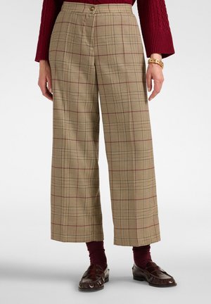 Person trägt beige karierte, weit geschnittene Hose, bordeauxroten Pullover, passende Socken, dunkelbraune Loafers und ein goldenes Armband an einem Handgelenk.