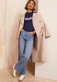 Beige trenchcoat päällä tummansinisellä T-paidalla, jossa on "Voilè"-printti, vaaleansiniset väljät farkut ja valkoiset tennarit, joissa on ruskeat yksityiskohdat.