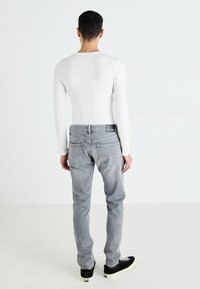 Camiseta blanca de manga larga, jeans ajustados grises, zapatillas negras con cordones blancos, con bolsillos traseros y etiqueta con logo en la cintura.
