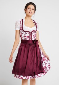 Jeune femme portant une robe dirndl florale blanche et bordeaux avec des manches en dentelle et un tablier bordeaux noué, debout devant un fond uni.