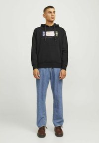 Black hooded sweatshirt with a front graphic, drawstrings, and a kangaroo pocket, paired with light blue loose-fitting jeans and brown shoes.
