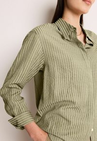 Chemise rayée verte avec un col boutonné et des manches longues, présentant un tissu texturé et des poignets retroussés.
