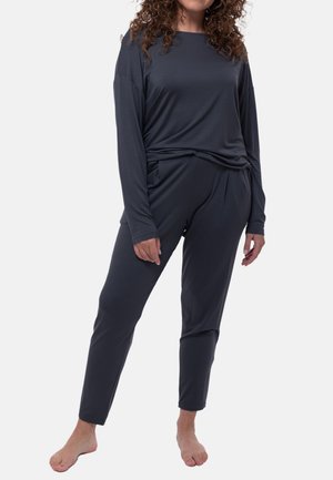 Vrouw staat blootsvoets, draagt een donker lang mouwen top en bijpassende taps toelopende broek tegen een effen lichte achtergrond.