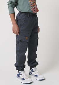 Temno sivi cargo hlače z elastiko na zapaklh, več žepi in bež aplikacijo; nosene z belimi in mornarsko modrimi supergami.