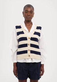 Triibuline kudumivest kreemjas ja tumesinises, millel on V-kaelus, nupulukk ja soonikuga äär, kantud valge kraega särgi ja tumesiniste lühikeste pükste peal.