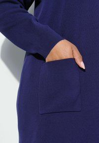 Kobaltově modré pletené šaty zblízka, s viditelnou rukou v kapse, s žebrovanou texturou a přiléhavými rukávy.