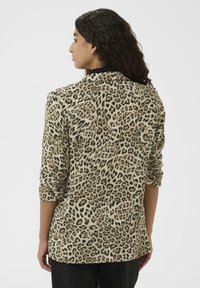 Moteris su ilgais plaukais, dėvinti smėlinį ir juodą leopardų rašto švarką, matoma iš nugaros prieš paprastą baltą foną.