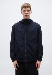 Tmavomodrá vetrovka s kapucňou, predným zipsom a pružnými manžetami. Vyznačuje sa hladkou, ľahkou textúrou a jemným švovým detailom.