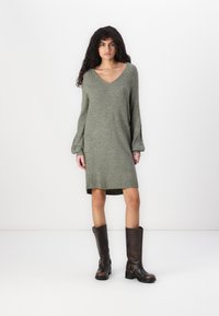 Robe en tricot vert olive avec un décolleté en V, des manches longues et une silhouette légèrement évasée, associée à des bottes marron montantes.