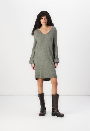 Abito a maglia verde oliva con scollatura a V, maniche lunghe e una silhouette leggermente svasata, abbinato a stivali marroni fino al ginocchio.