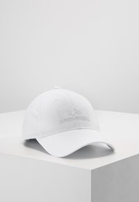 Bjela bejzbol kapa s zakrivljenim vizirjem, izrađena od tkanine, sa teksturiranim zakrpom na prednjoj strani koji sadrži tekst "ALPHA INDUSTRIES" i logo.