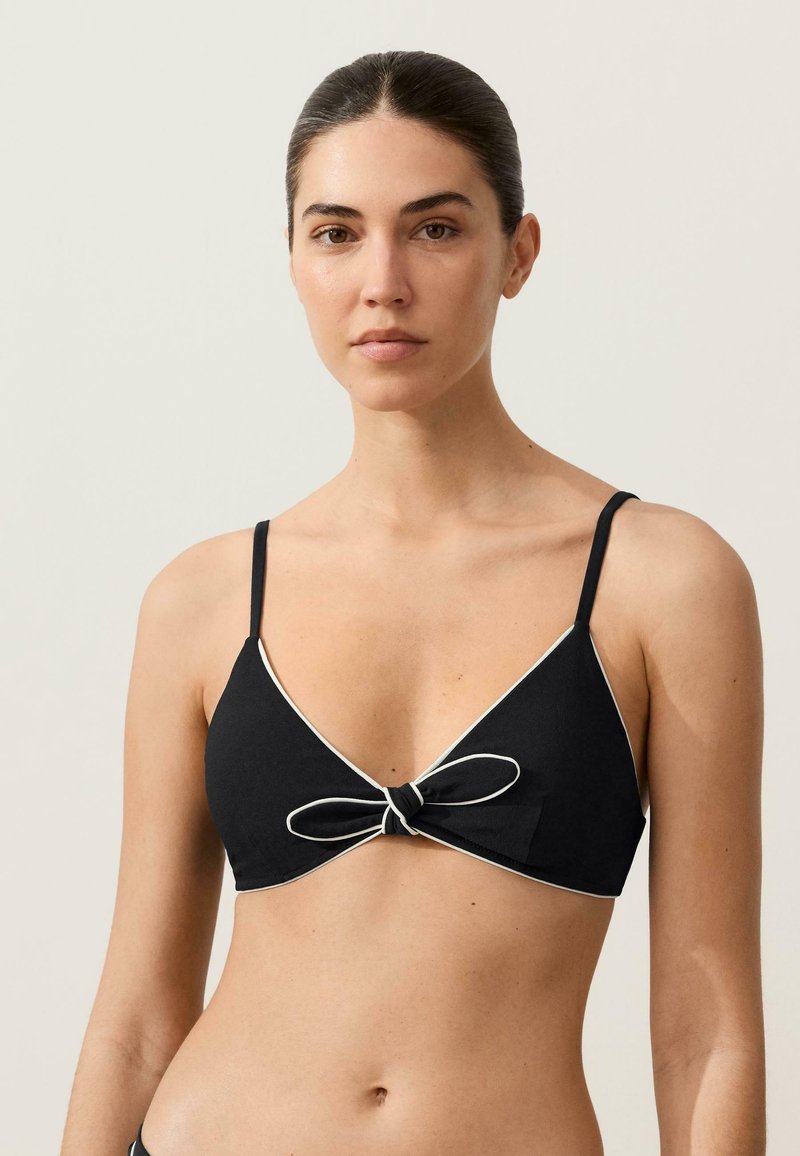 Kvinde med mørkt hår iført en sort bikini-top med sløjfe foran og hvid kant, stående mod en ensartet lys baggrund.