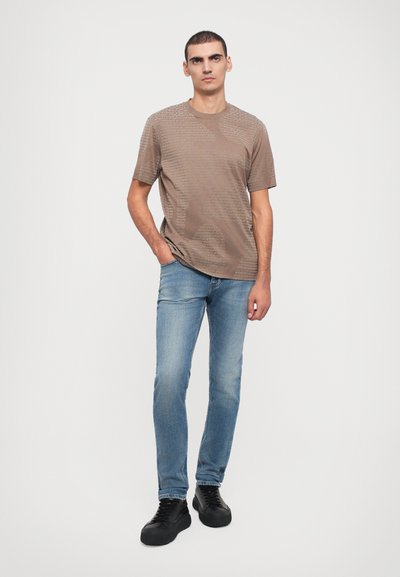 Brun t-skjorte med kort erme og teksturert mønster, kombinert med lyseblå jeans og svarte snøresko. Modellen står foran en nøytral bakgrunn.