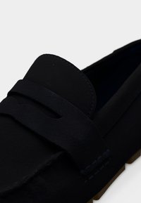 Μαύρο μοκασίνι από σουέντ με σχέδιο moc toe, λεπτομέρεια με λουράκι penny και καφέ ελαστική σόλα σε λευκό φόντο.