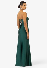Abito verde smeraldo senza spalline con un design fluido. Presenta una schiena laccata e un tessuto morbido e liscio. Silhouette elegante con texture drappeggiata.
