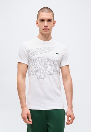 Jeune homme aux cheveux courts portant un t-shirt blanc avec le contour noir d'un crocodile et un petit logo vert sur la poitrine, debout devant un fond uni.