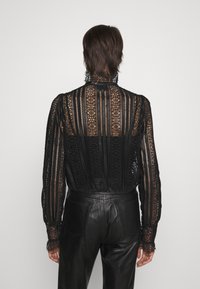 Blouse en dentelle noire avec motifs floraux et rayures verticales, à col haut et manches longues, associée à un pantalon en cuir noir.