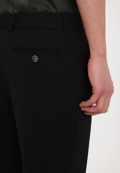 Μαύρο κομμένο παντελόνι διαθέτει επίπεδη τσέπη με διακοσμητικό κουμπί. Το ύφασμα φαίνεται λείο και καλοσχηματισμένο. Ένα χέρι είναι τοποθετημένο κοντά στη μέση.