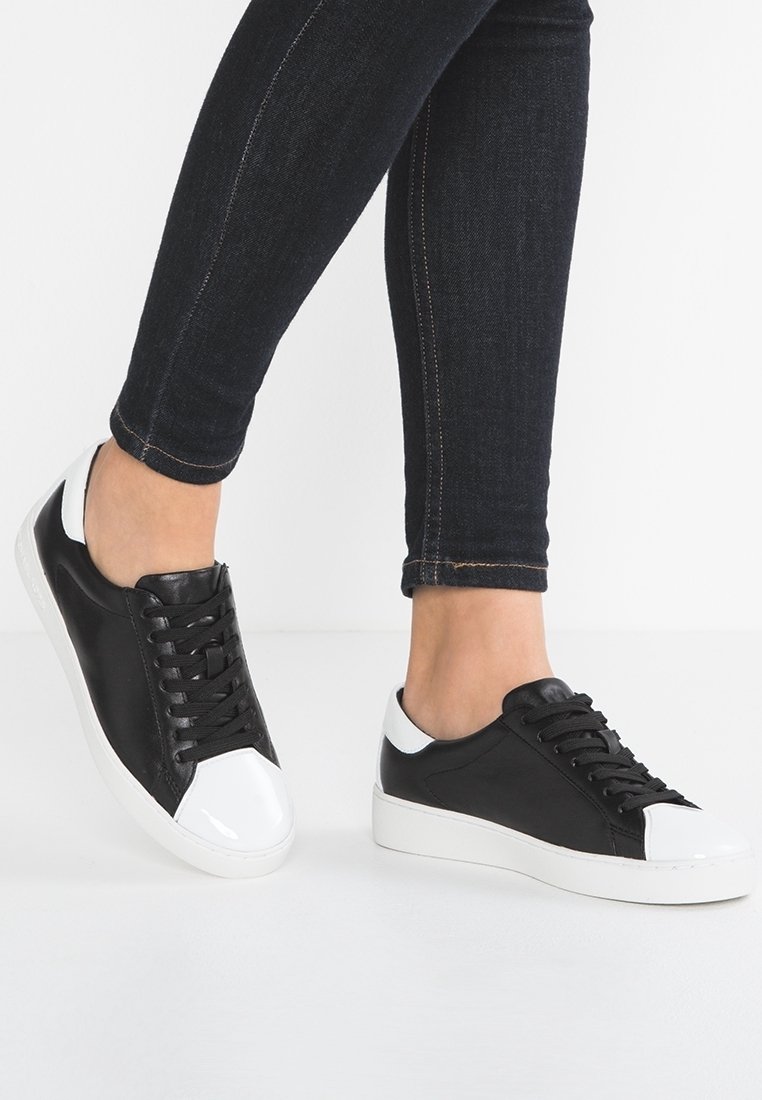 Jambes en jean skinny foncé portant des baskets basses noires et blanches avec des semelles blanches sur un fond clair uni.