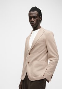 Άνδρας που φοράει μπεζ σακάκι πάνω από λευκό πουκάμισο και σκούρο παντελόνι, στέκεται με το ένα χέρι στην τσέπη μπροστά σε απλό φωτεινό φόντο.