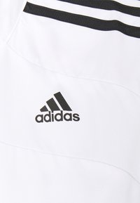 Camisa deportiva blanca con tejido texturizado, diseño de tres franjas negras en la parte superior y un logo negro de Adidas en el pecho.