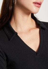 Svart V-ringad topp med en strukturerad krage, slät textur och figurnära design, som visar en närbild av halsringningen och kragdetaljer.