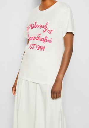 Nainen, jolla on valkoinen T-paita, jossa vaaleanpunaista kursiivitekstiä "Philosophy di Lorenzo Serafini EST. 1984", ja valkoinen hame.