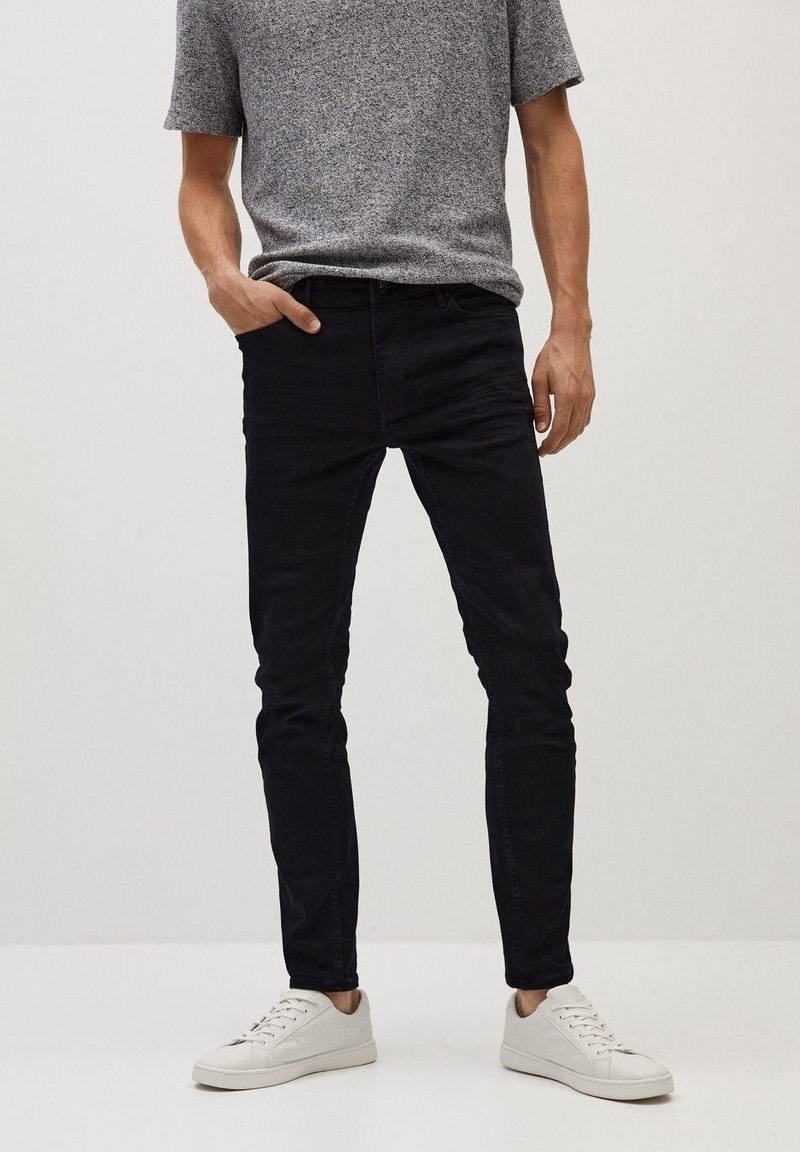 Homme portant un jean noir slim, une chemise grise à manches courtes et des baskets blanches, debout avec une main dans la poche devant un fond uni.