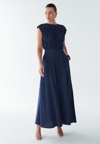 Tamsiai mėlyna maksi suknelė su prigludusiu liemeniu, klostėtu dizainu ir trumpomis rankovėmis. Lygus audinys, tekanti suknelės apačia ir subtilus blizgesys.