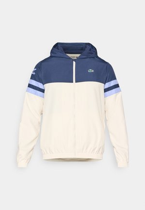 Veste à capuche en crème, marine et bleu clair avec fermeture éclair, poignets élastiques et logo Lacoste sur la poitrine. Présente un design coloré en blocs.