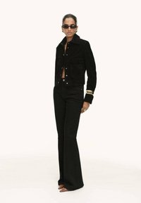Giacca in corduroy nera con tasche anteriori, abbinata a pantaloni neri a vita alta e svasati. Il modello indossa occhiali da sole rotondi e accessori dorati.